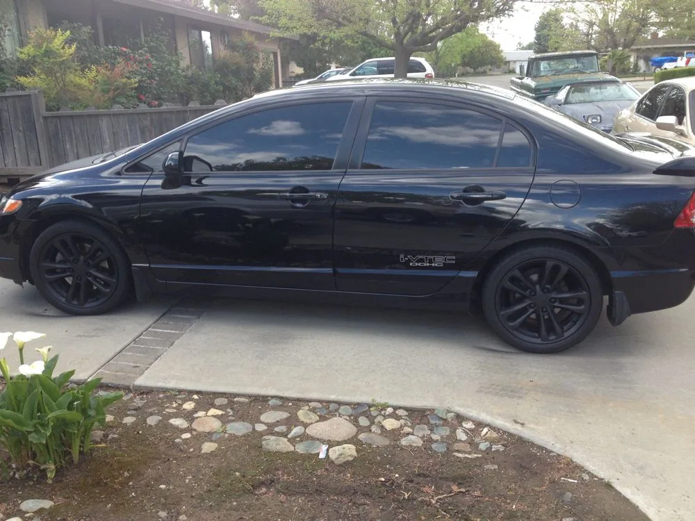 Black sedan with tinted windows parked in driveway with landscaping and trees visible