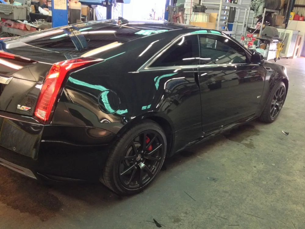 Black coupe with tinted windows in an automotive workshop, showing side profile with red taillights and dark wheels